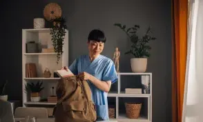 Asian dedicated nurse in blue scrubs is seen preparing medical tools and organizing documents into a brown backpack in a well-lit, modern room with shelves and assorted decor.