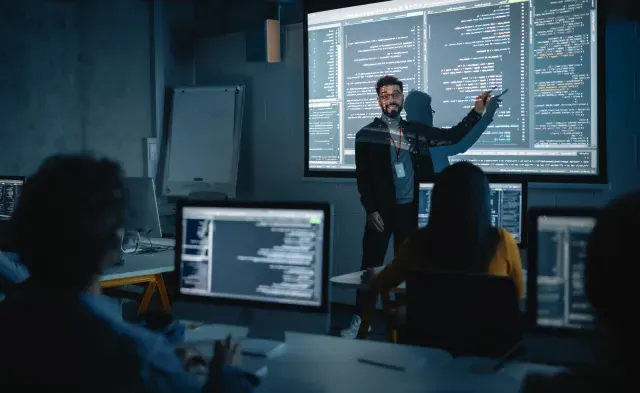 Teacher Giving Computer Science Lecture to Diverse Multiethnic Group of Female and Male Students in Dark College Room. Projecting Slideshow with Programming Code. Explaining Information Technology.